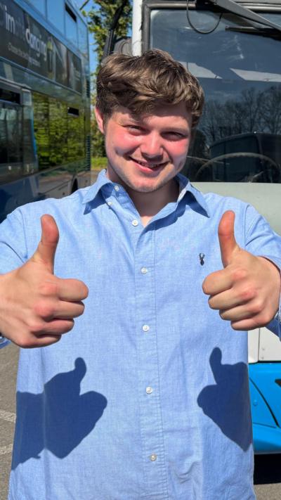 Luke Wilson puts two thumbs up as he stands in front of two, parked Ulsterbuses. He is a young man with short, light coloured hair and a blue shirt.