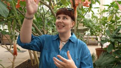 Matilda Brown has a dark red fringe and is wearing a blue shirt with glasses on her head. She has one hand up and is looking towards her hand while smiling