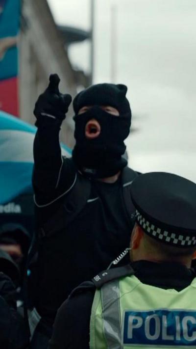 Person in balaclava pointing and shouting as a police officer watches on