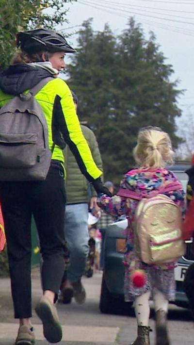 A parent wearing cycling safety gear holding the hand of a child whilst walking to school