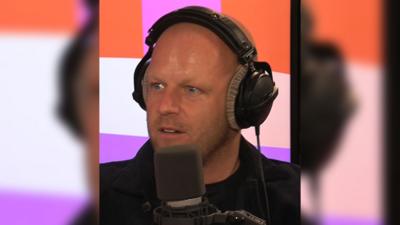 Man with stubble, wearing headphones talks into a mic in a radio studio.