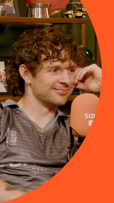Tom Grennan smiles - he has short curly brown hair and wears a grey top