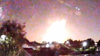 A bright flash in the night sky over some houses and gardens.