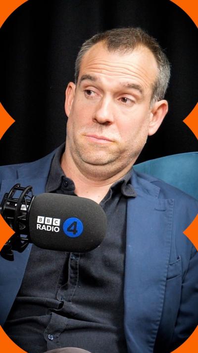 A man with short hair wearing a black shirt under a blue blazer raises his eyebrows at someone off-camera