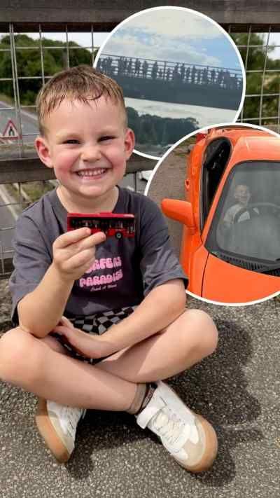 A young boy sits cross legged, smiling to camera while holding a red bus toy. A road can be seen in the background.