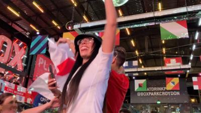 A woman wearing an England shirt and bucket hat waving her arm in the air and sticking out her tongue in celebration
