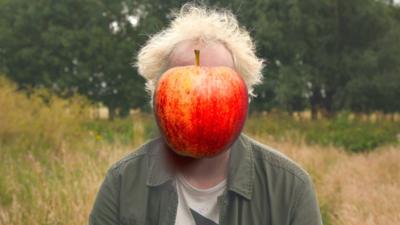 A person with messy blonde hair has their face obstructed by a large apple