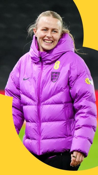 Hannah Hampton smiling, wearing a purple puffa jacket on the pitch.