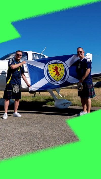 Two men stand beside a small plane, holding a saltire.