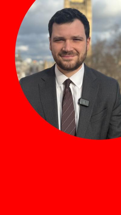 Henry Zeffman wears a suit and tie, and stands outside with Westminster in the background 