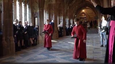 Two boys in red cassocks holding frying pans in a tiled corridor with children lining the sides of it