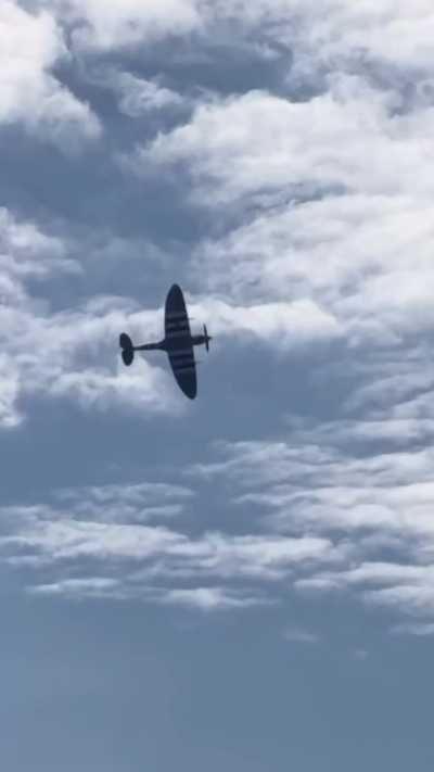 A Spitfire aircraft mid-flight in blue sky with clouds in the background.