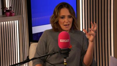 Woman with short brown hair, wearing a dark grey knit jumper. She is speaking in to a pink microphone with SCOTCAST written on it.