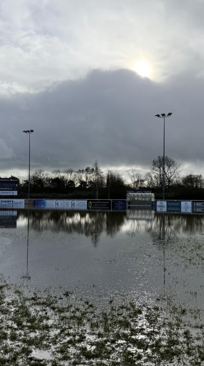 Flood water is seen on the pitch at Taunton Rugby Club under grey skies. In the background are advertising hoardings and two floodlights