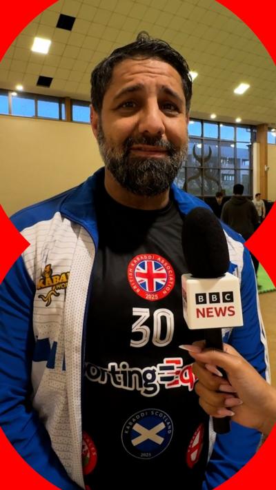 A middle-aged man looks directly at the camera. He's dressed in a blue and white jacket and black t shirt. Someone is holding a BBC News branded microphone under his mouth.