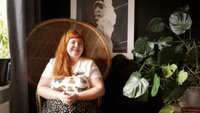 A woman with long red hair is smiling and sitting in a brown armchair. There is a plant to her left and a poster of Freddie Mercury behind her.