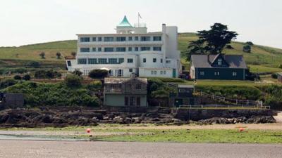 Burgh Island Hotel. White with a green top of it. A smaller dark building sits to the right and a smaller building again further down the hill in front of the hotel is also visible. The hill continues up behind the hotel.