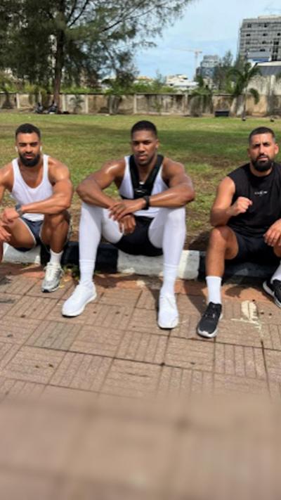Anthony Joshua sits between Sina Ghami and Latif Ayodele as all wear training kit, with a grassy area behind the three men