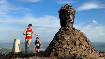 Tow men in shorts and race tops run past a cairn on top of a hill against a blue sky.