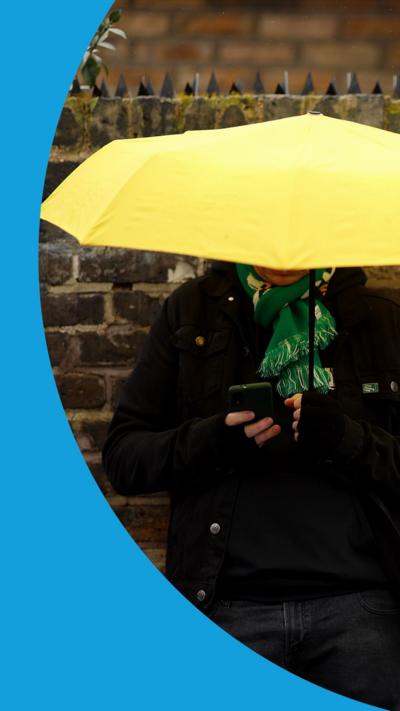A man wearing a black jacket and jeans, and a green football scarf is using his phone and sheltering under a yellow umbrella.