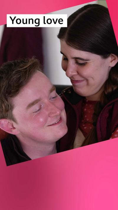 A woman sits on a man's knee, both with coy smiles on their faces