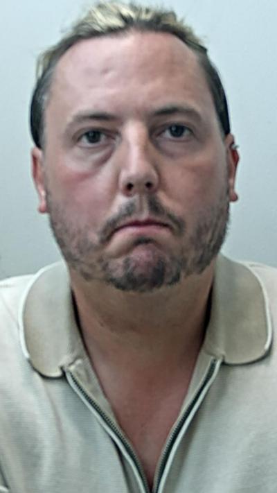 mugshot of man in police custody wearing polo top. He has a beard and long blonde hair.