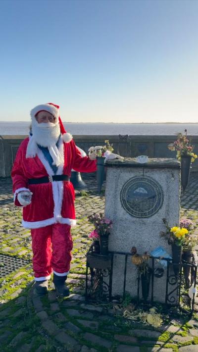Man dressed in a bright red Santa Claus costume standing beside a stone memorial adorned with flowers and plaques. The setting is an outdoor waterfront area with cobblestone ground, a fishing rod leaning against the wall, and a clear view of the water under a bright sky.