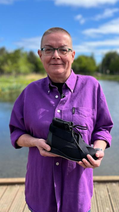 Jeanette Russell smiles and she stands in front of a body of water. It's a sunny day and there is lush green grass on either side with a row of trees and hedges on either side. She has cropped hair and is wearing glasses and a bright purple shirt. She is holding the black lace-up shoe