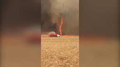 Flames in the shape of a tornado on a field, with a truck spraying water on it