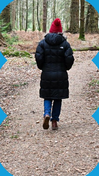 A person seen from behind walking along a walking trail in a forest. They have a long, black padded coat on.