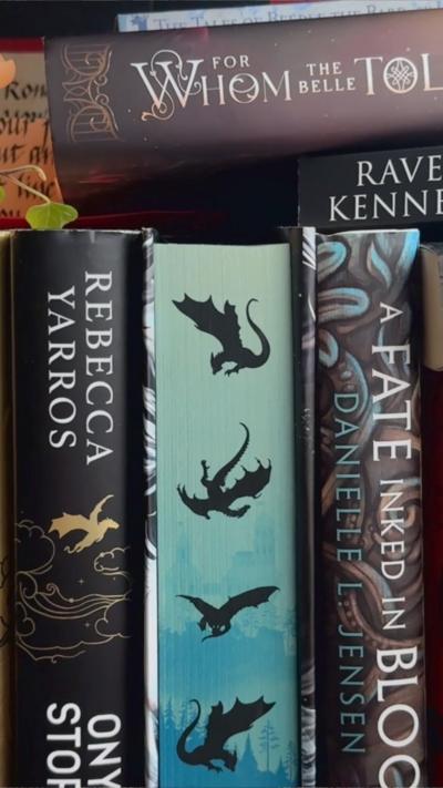 Books lined up on a shelf, one is a Rebecca Yarro, another is a For Whom the Belle Toll book and other book binds have mythical creatures with blue and purple colours. 