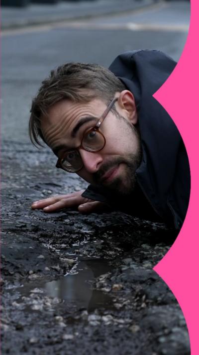 A man in a black coat lies on a road next to a pothole that has water in it.