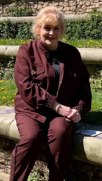 Lucy Ashton sits looking at the camera. She is wearing a dark red jacket and matching trousers.