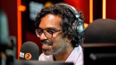 Romesh Ranganathan wears headphones while speaking into a microphone in a BBC Radio 2 studio.