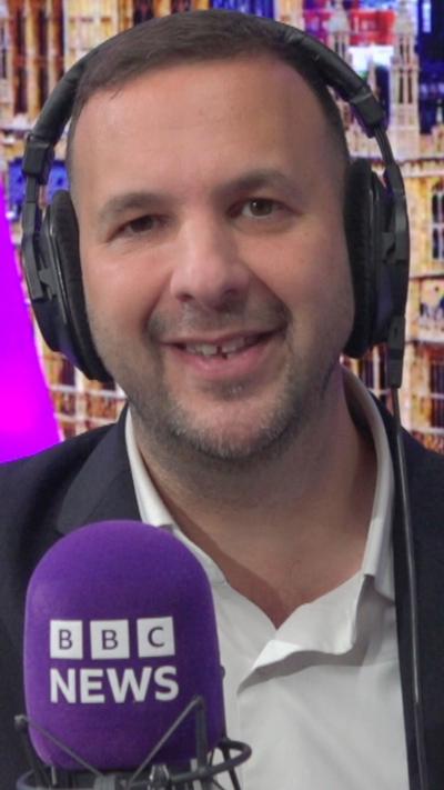 Green Party leader Zack Polanski wears a suit jacket and wears headphones as he speaks into a purple BBC microphone