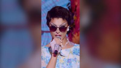 Drag queen Bones wears a floral dress with a short dark wig and sunglasses. She holds a sparkly mic