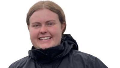 Councillor Grace Lewis is wearing a black waterproof coat with a hood and smiling. She has fair wavy hair, tied back. She is in front of a white background.