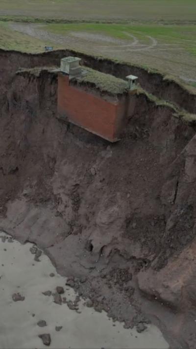 A coastal cliff with sandy beach at the bottom and a rectangular red brick building with three chimneys exposed in the cliff -face. 