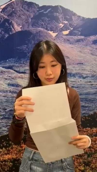 Pupil with long hair, wearing brown top and jeans standing in front of mountain wallpaper opens an A4 envelope.