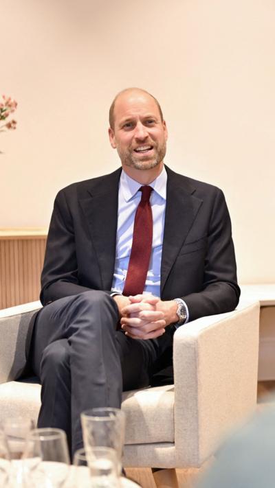 a balding man with a beard sat in a white chair. He is wearing a blue blaze, grey trousers, blue shirt and claret tie