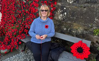 A woman sitting on a bench, wearing sunglasses, a blue jumper. She is surrounded by crocheted poppies