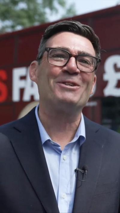 Andy Burnham, wearing a dark grey jacket over a light blue shirt, smiles at the camera. He has dark short hair and is wearing glasses