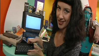 Gia Milinovich staring into the camera holding a Sinclair ZX Spectrum, with other computers in the background.