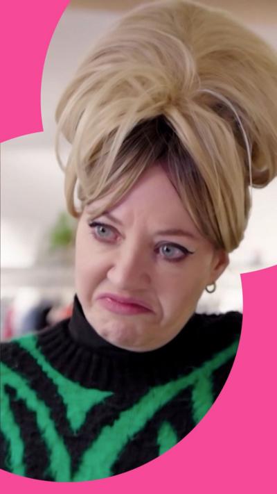 Character Mandy pulls a face in a shop - she has a blonde beehive hairstyle and wears a green and black jumper 