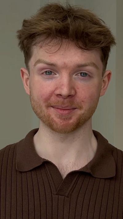 A man in a burgundy polo-shirt is looking directly into the camera and smiling. He has dark-coloured hair and a light beard. 