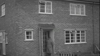 A man opening the front door of a semi-detached brick house.