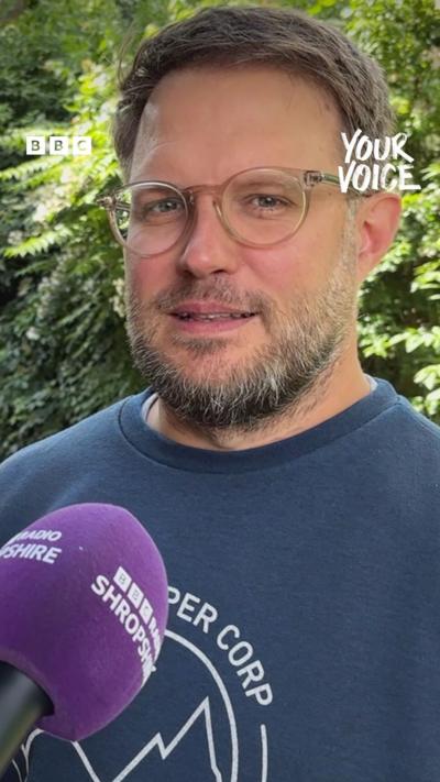 A man with brown hair and beard with glasses and a blue top talking into a purple microphone