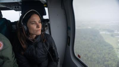 A woman, Ione Wells, looks out of the window in a helicopter