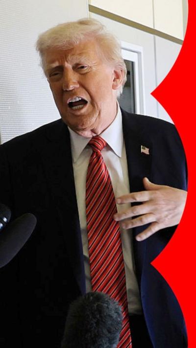 Donald Trump, wearing a red tie, looks to the side in front of microphones