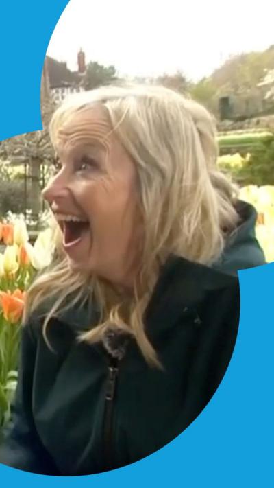 Carol Kirkwood wears a green coat and smiles in surprise against a backdrop of yellow and orange flowers